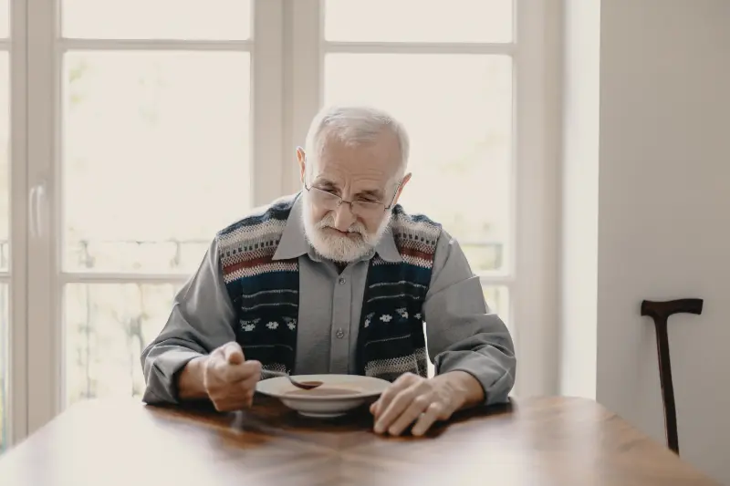 An elderly man suffering from metallic taste due to his recently starting cancer treatment is attempting to eat soup.